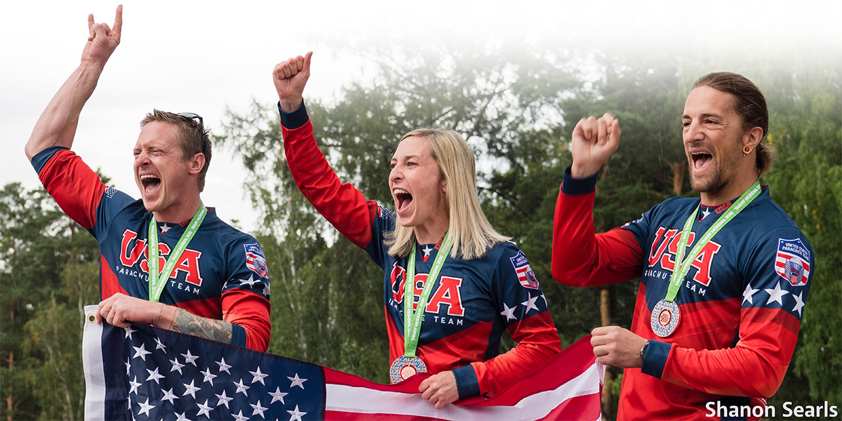 Three members of the U.S. Parachute Team wearing medals, appearing as shouting, holding an American Flag in one hand and pumping their other hands in the air